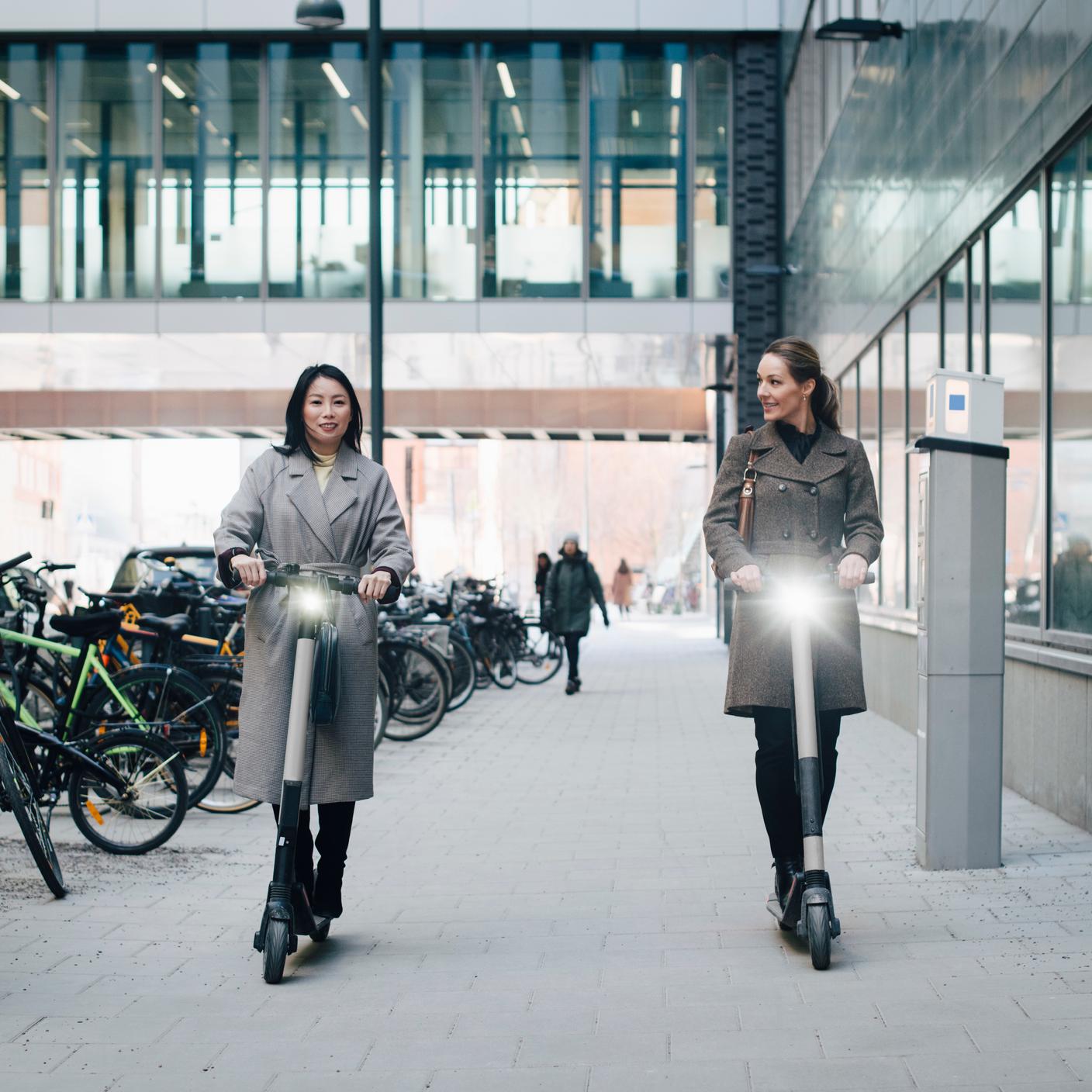 Female colleagues riding electric push scooter on footpath by building in city