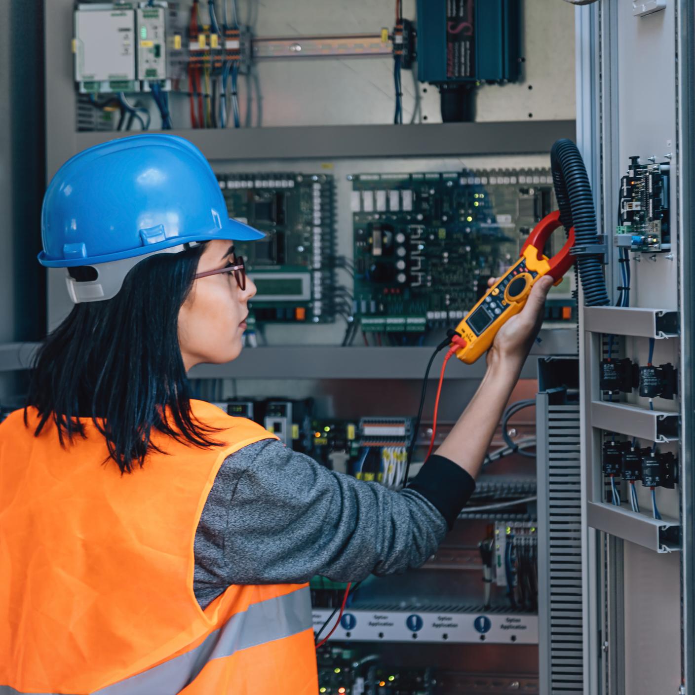 Young woman industrial service electrician engineer wearing protective vest and blue technician helmet testing and checking fridge electric voltage.